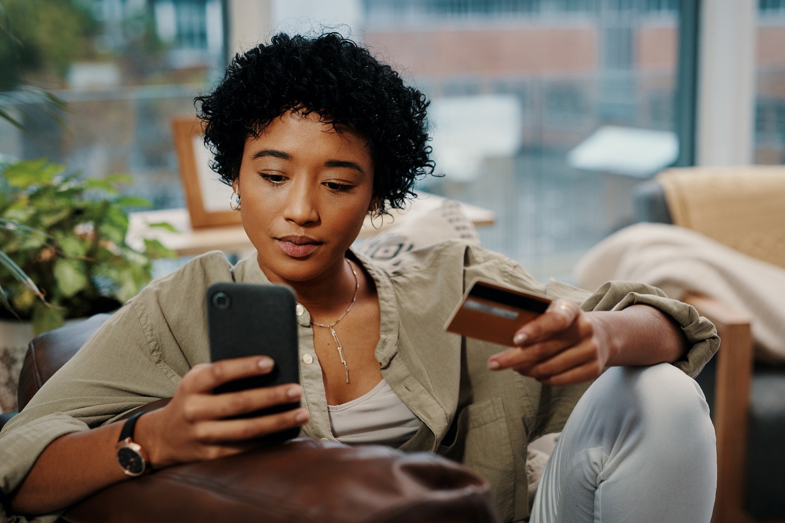 Mulher negra sentada no sofá com o celular em uma mão e um cartão de crédito na outra fazendo compras online